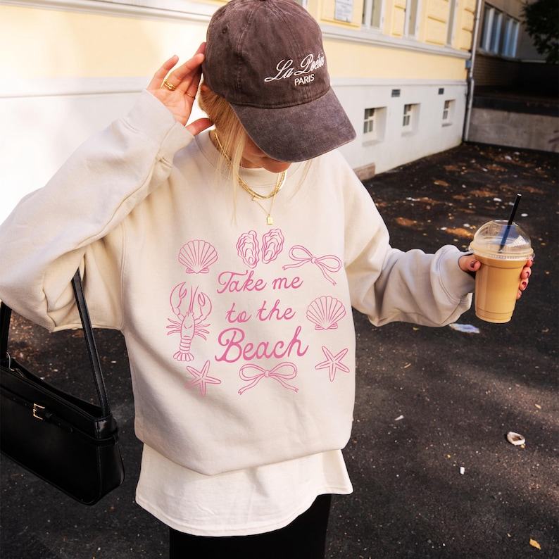May include: A cream-colored sweatshirt with the phrase "Take me to the Beach" in pink, along with seashell and bow graphics. A brown baseball cap with "La Petite Paris" text is worn.
