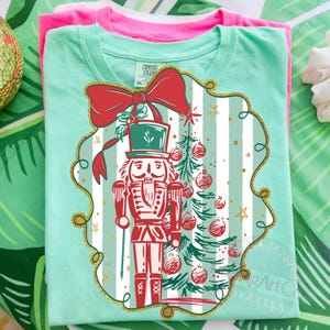 May include: A folded, light green t-shirt with a Christmas-themed graphic. The graphic features a red nutcracker soldier, a Christmas tree, and a red bow. The design is framed with gold glitter accents. A pink t-shirt is visible underneath.