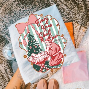 Puede incluir: Camiseta azul claro con un dise&ntilde;o navide&ntilde;o. El dise&ntilde;o incluye un lazo rosa, un &aacute;rbol de Navidad y el texto "Every Knee Shall Bow" con una representaci&oacute;n de Pap&aacute; Noel arrodillado. La camiseta tiene un tema festivo y religioso.
