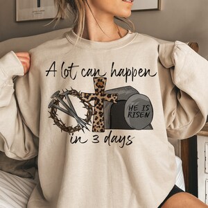 May include: A beige sweatshirt with a graphic print of a cross with a leopard print design, nails, a crown of thorns, and a stone with the text "He is Risen". The text "A lot can happen in 3 days" is printed on the sweatshirt.