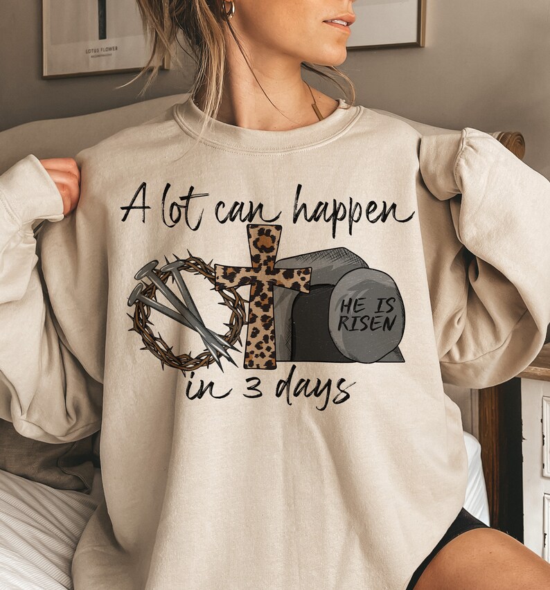 May include: A beige sweatshirt with a graphic print of a cross with a leopard print design, nails, a crown of thorns, and a stone with the text "He is Risen". The text "A lot can happen in 3 days" is printed on the sweatshirt.