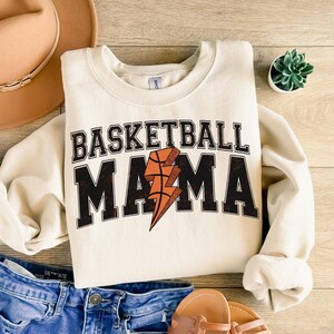 May include: A cream-colored sweatshirt with the text "Basketball Mama" in black and orange lettering. A lightning bolt with a basketball inside is featured in the center of the text.