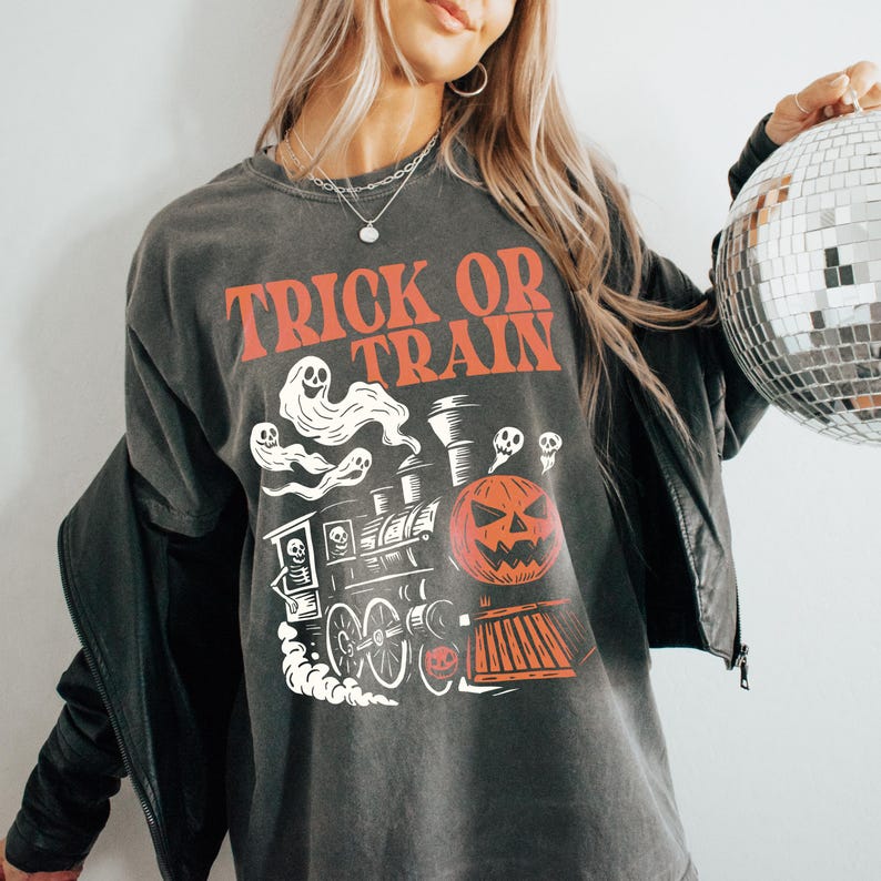 May include: Dark grey t-shirt with the text "TRICK OR TRAIN" in orange. The graphic features a train with a jack-o'-lantern, ghosts, and a skeleton. A fun Halloween-themed tee.