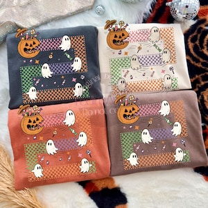 May include: Four sweatshirts in various colors, each featuring a Halloween-themed design. The design includes a jack-o'-lantern with candy, ghosts, and candy. The background has a checkered pattern and horizontal stripes in different colors.