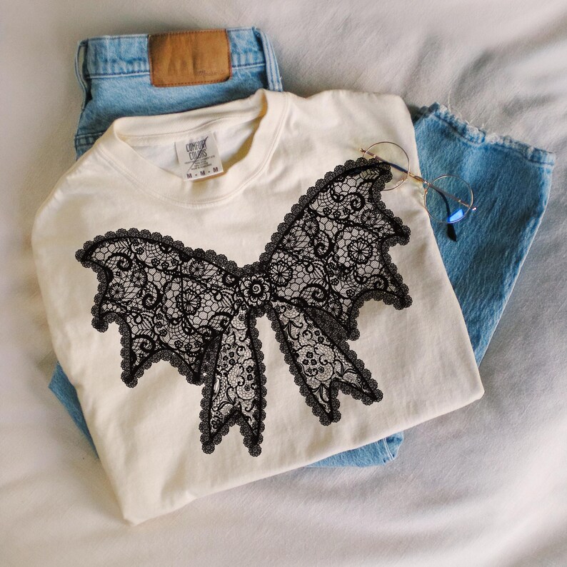 May include: Cream-colored t-shirt featuring a large black lace bow design. The shirt is paired with light blue denim jeans. The bow design is intricate, with detailed floral patterns. A pair of round glasses rests on the jeans.