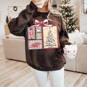 May include: A dark brown sweatshirt with a festive Christmas design. The design features a large gift box with a red bow, and inside are postage stamp-style images of a nutcracker, Santa, a Christmas tree, and a sleigh.