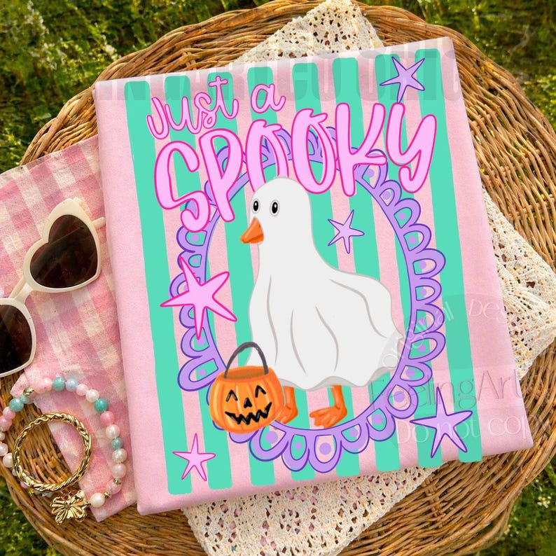 May include: A light pink t-shirt with teal vertical stripes featuring a white ghost wearing a sheet, holding an orange jack-o'-lantern, surrounded by a purple scalloped oval frame and pink stars, with the text Just a SPOOKY printed above.