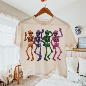 Puede incluir: Una camiseta color crema con un gr&aacute;fico colorido de cuatro esqueletos bailando. Cada esqueleto es de un color diferente: naranja, morado, verde y rosa. Los esqueletos llevan gafas de sol y saludan.