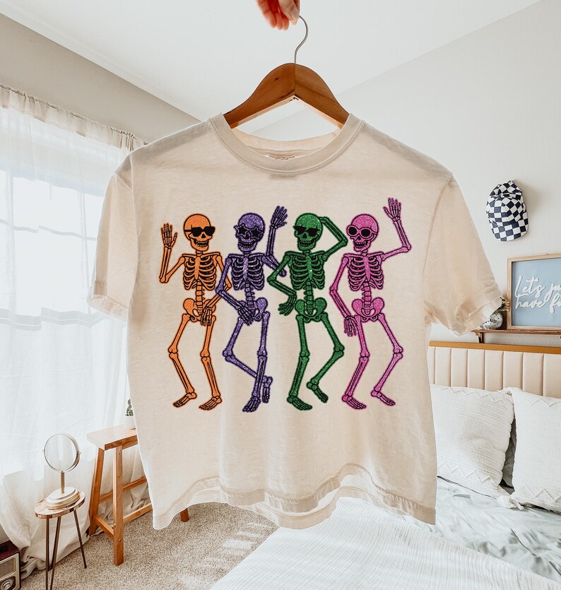Puede incluir: Una camiseta color crema con un gr&aacute;fico colorido de cuatro esqueletos bailando. Cada esqueleto es de un color diferente: naranja, morado, verde y rosa. Los esqueletos llevan gafas de sol y saludan.