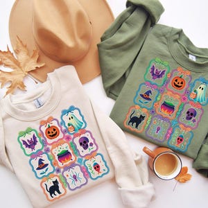 May include: Two sweatshirts, one cream and one olive green, each featuring a colorful Halloween-themed design. The design includes a bat, jack-o'-lantern, ghost, witch hat, cauldron, skull, spider, and a black cat. A tan hat and a fall leaf are also in the image.