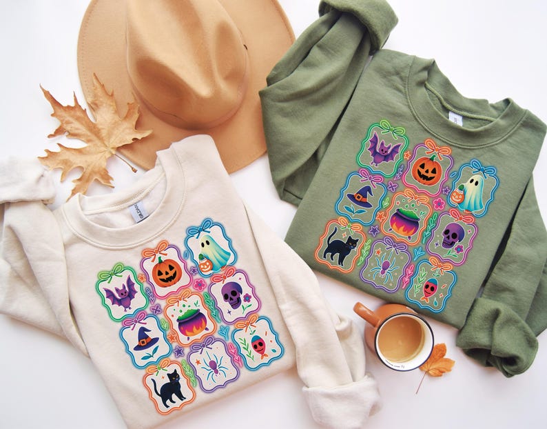 May include: Two sweatshirts, one cream and one olive green, each featuring a colorful Halloween-themed design. The design includes a bat, jack-o'-lantern, ghost, witch hat, cauldron, skull, spider, and a black cat. A tan hat and a fall leaf are also in the image.