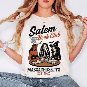 May include: Cream-colored t-shirt featuring the text "Salem Book Club" with illustrations of three witches reading books. The design includes bats and the words "Massachusetts Est. 1692."