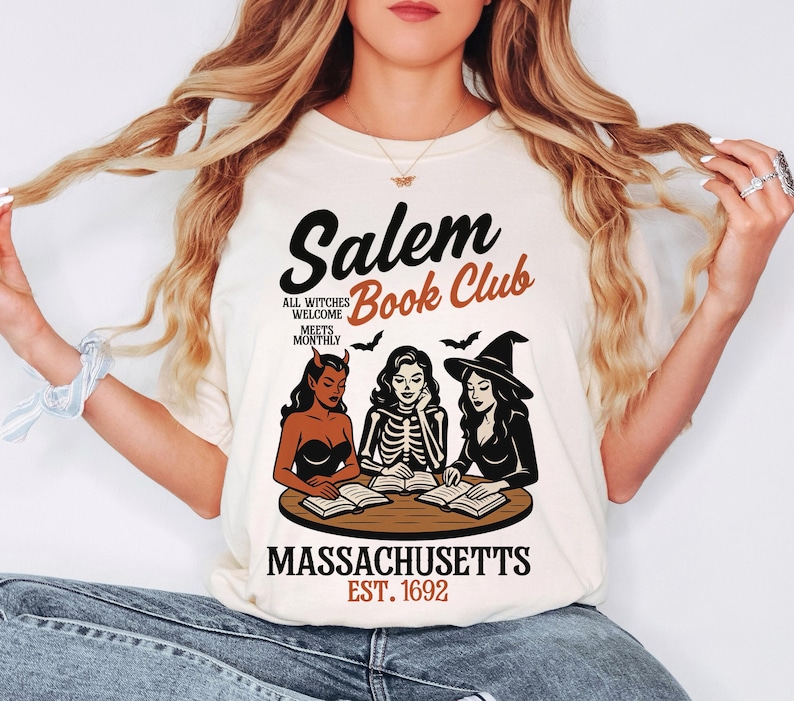 May include: Cream-colored t-shirt featuring the text "Salem Book Club" with illustrations of three witches reading books. The design includes bats and the words "Massachusetts Est. 1692."