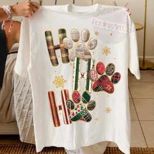 K&ouml;nnte beinhalten: Wei&szlig;es T-Shirt mit festlichem Design, das die Worte "HO HO HO" und Pfotenabdr&uuml;cke zeigt. Die Buchstaben und Pfotenabdr&uuml;cke sind mit Karomustern, Blumenmustern und Schneeflocken in Rot, Gr&uuml;n und Gold verziert. Ein grafisches T-Shirt zum Thema Feiertage.