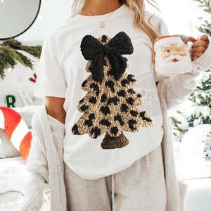 May include: White t-shirt featuring a knitted Christmas tree design. The tree has a leopard print pattern in tan and black, topped with a black bow. The tree is a festive and unique holiday design.