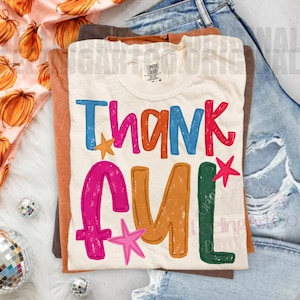 May include: A cream-colored t-shirt featuring the word THANKFUL in large, multi-colored, distressed-style letters. The text is printed in blue, pink, orange, and green, accented with small pink and orange stars. A Comfort Colors brand tag is visible.