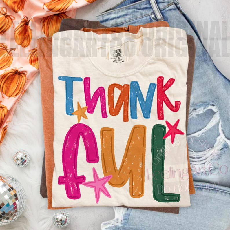 May include: A cream-colored t-shirt featuring the word THANKFUL in large, multi-colored, distressed-style letters. The text is printed in blue, pink, orange, and green, accented with small pink and orange stars. A Comfort Colors brand tag is visible.