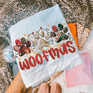 Puede incluir: Camiseta azul claro con el texto "Merry Woofmas" en rojo, con un dise&ntilde;o de huella de pata con luces navide&ntilde;as y patrones festivos. El dise&ntilde;o incluye detalles dorados y una mezcla de texturas.