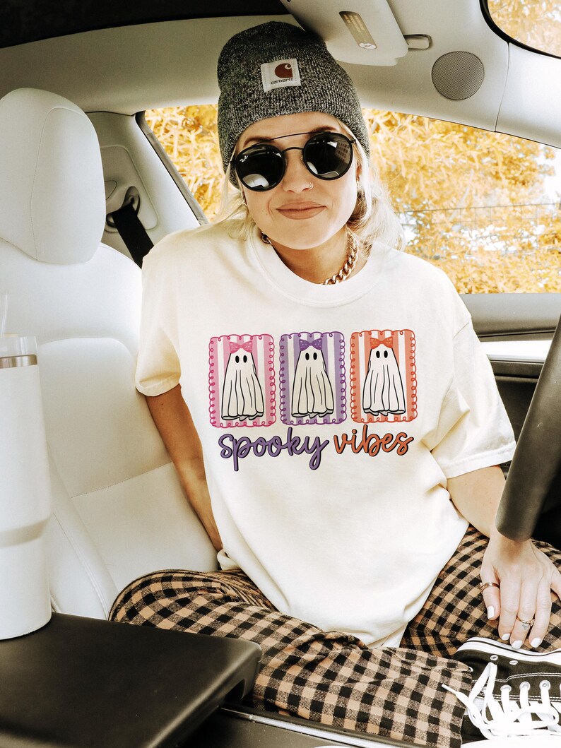 May include: Cream-colored t-shirt with a Halloween-themed design. The shirt features three ghost illustrations within decorative frames, with the words "spooky vibes" in a script font. The person in the image is wearing a gray beanie and sunglasses.