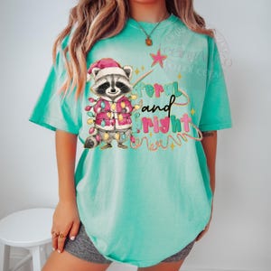 May include: A turquoise t-shirt features a cartoon raccoon wearing a Santa hat and a pink coat adorned with Christmas lights. The text "feral and bright" is written in a playful font, with a star and decorative lines.