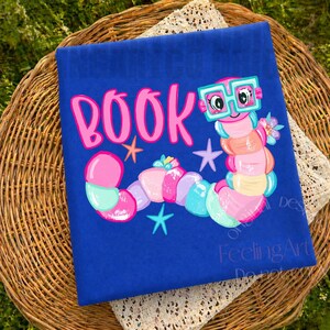 May include: A royal blue t-shirt featuring a colorful graphic of a segmented worm wearing teal-rimmed glasses. The word BOOK is printed in large pink letters above the worm, accompanied by small teal and orange star shapes and a flower.