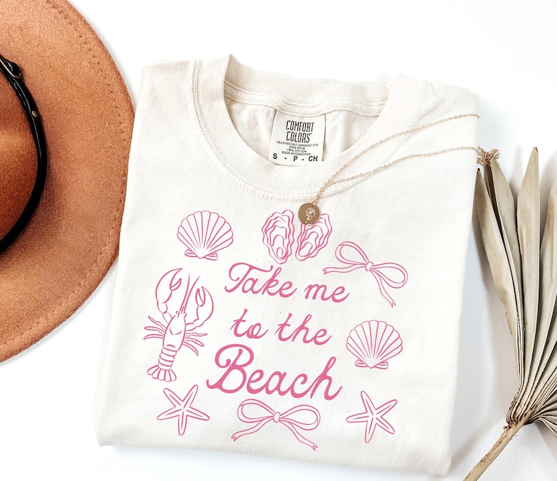 May include: Cream-colored t-shirt with pink illustrations of seashells, a lobster, starfish, and bows. The text "Take me to the Beach" is written in a cursive font. A gold necklace and a brown hat are also visible.