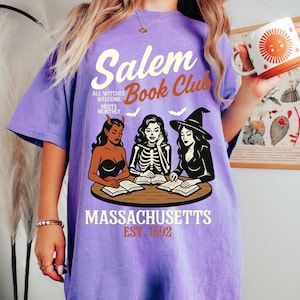 May include: A purple t-shirt with the text "Salem Book Club" and an illustration of three witches reading books. The shirt also includes the text "All witches welcome meets monthly" and "Massachusetts Est. 1692".