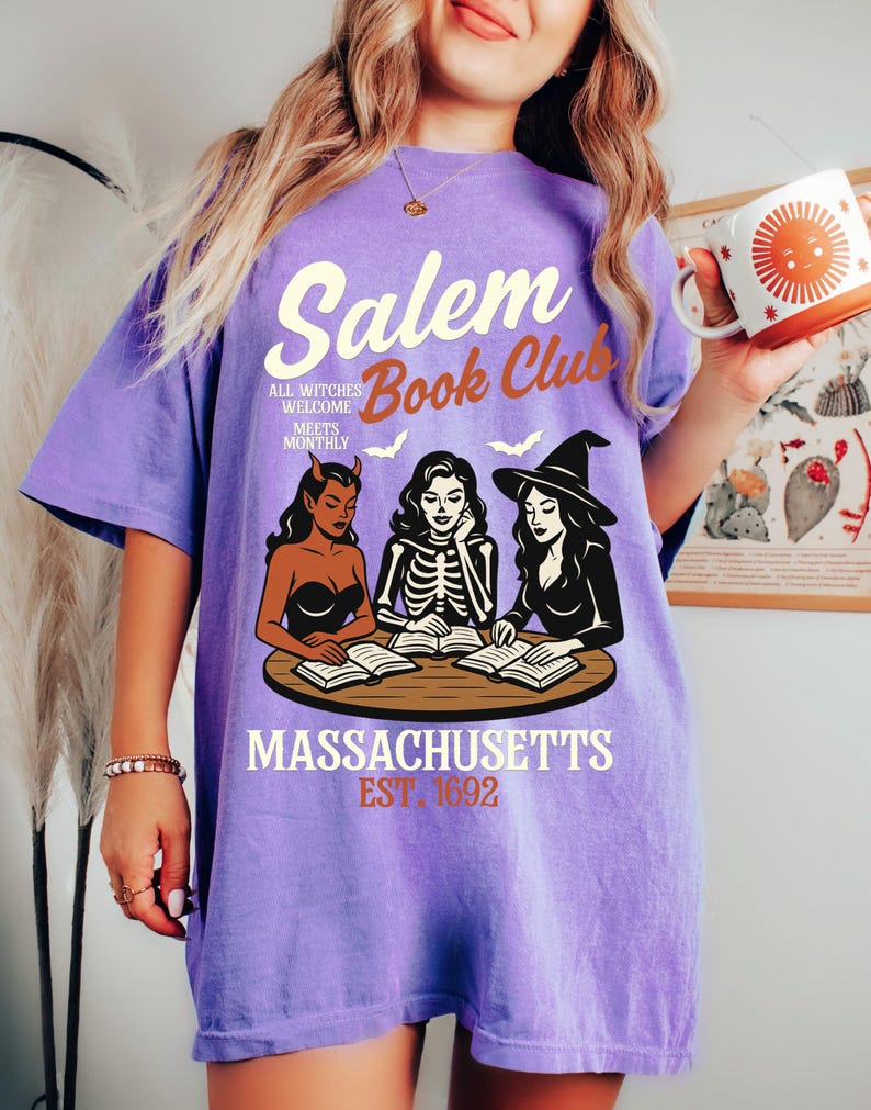 May include: A purple t-shirt with the text "Salem Book Club" and an illustration of three witches reading books. The shirt also includes the text "All witches welcome meets monthly" and "Massachusetts Est. 1692".