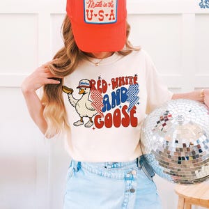 May include: A person wearing a red trucker hat with a "Made in the U.S.A." patch and a cream-coloured t-shirt. The t-shirt features a cartoon goose holding a hot dog, with the text "Red, White, and Goose" in red, white, and blue.