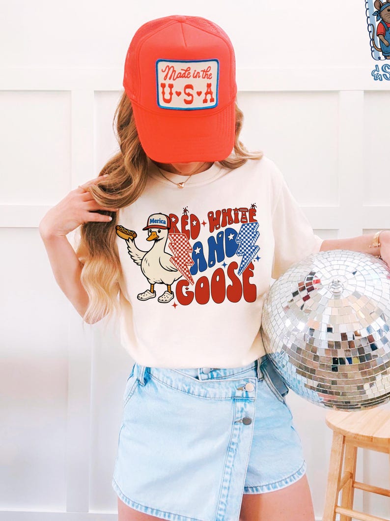 May include: A person wearing a red trucker hat with a "Made in the U.S.A." patch and a cream-coloured t-shirt. The t-shirt features a cartoon goose holding a hot dog, with the text "Red, White, and Goose" in red, white, and blue.