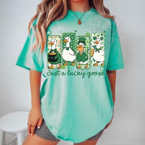May include: A turquoise t-shirt featuring a graphic of four cartoon geese in St. Patrick's Day attire. Each goose is in a rectangular frame with shamrocks and rainbows. The text "Just a lucky goose" is below the geese.