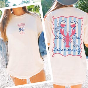 Puede incluir: Camiseta color crema con un diseño de copa de cóctel rosa y azul en la parte delantera. La parte trasera presenta una langosta, copas de vino y el texto "Cin Cin Dolce vibes only" en rosa y azul.