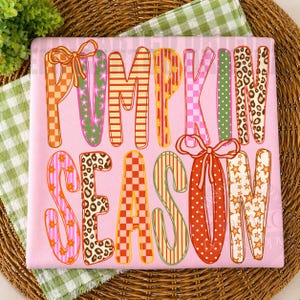 May include: A light pink fabric square featuring the text PUMPKIN SEASON in large, multi-patterned letters. The letters display various designs including orange and white checkers, green stars, leopard print, orange stripes, and polka dots.