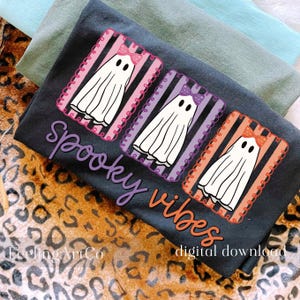 May include: A dark gray t-shirt featuring three cartoon ghosts with bows, set against striped backgrounds of pink, purple, and orange. The words "Spooky vibes" are written in purple and orange script. The phrase "digital download" is also visible.