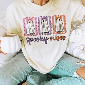 May include: Cream-colored t-shirt with a Halloween theme. The shirt features three cartoon ghosts with bows in pink, purple, and orange frames. The words "Spooky Vibes" are written in a purple script font below the ghosts.