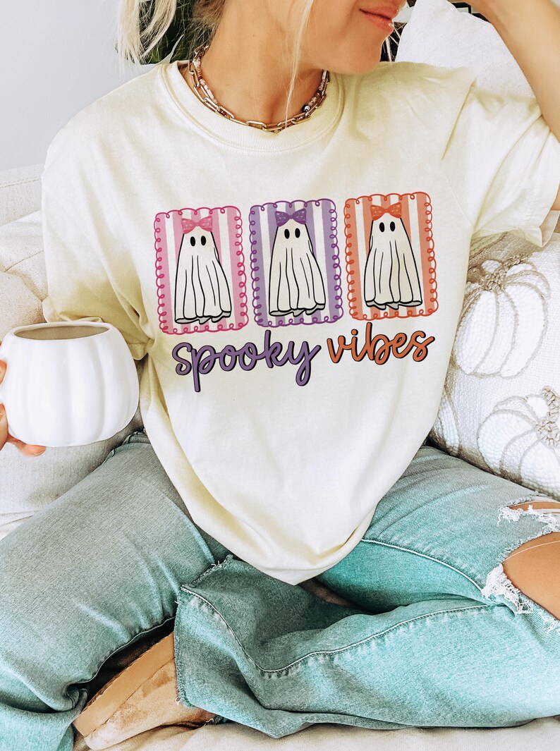 May include: Cream-colored t-shirt with a Halloween theme. The shirt features three cartoon ghosts with bows in pink, purple, and orange frames. The words "Spooky Vibes" are written in a purple script font below the ghosts.
