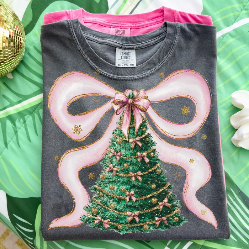 Pu&ograve; includere: Due magliette sovrapposte, una rosa e una grigio antracite. La maglietta superiore presenta un grande design a fiocco rosa con un albero di Natale al centro. L'albero &egrave; verde con fiocchi rosa e dettagli dorati. La maglietta grigia ha il testo "Comfort Colors".