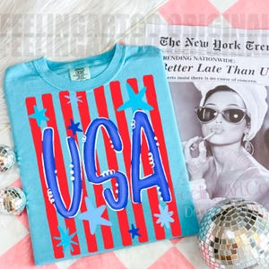 May include: A light blue t-shirt with a patriotic design. The shirt features red vertical stripes, blue stars, and the word "USA" in a bold, blue font. A casual top for celebrating.