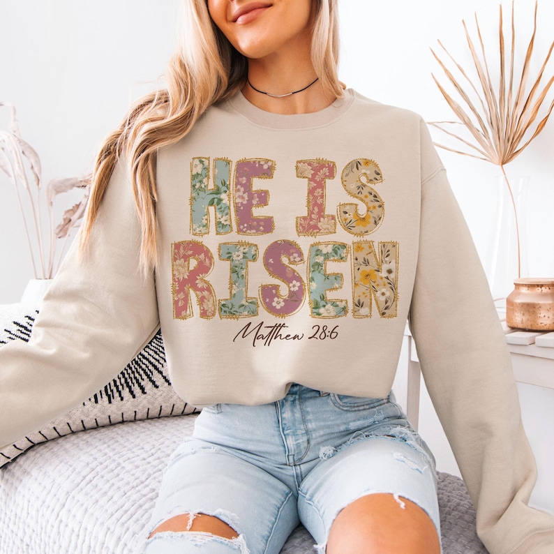 May include: A beige sweatshirt with a floral print design that reads "He is Risen" with the scripture reference "Matthew 28:6" underneath.