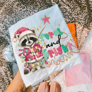 May include: Light blue t-shirt featuring a raccoon wearing a Santa hat and a pink coat adorned with Christmas lights. The text "feral and bright" is written in colorful, playful lettering. A pink star and gold stars complete the design.