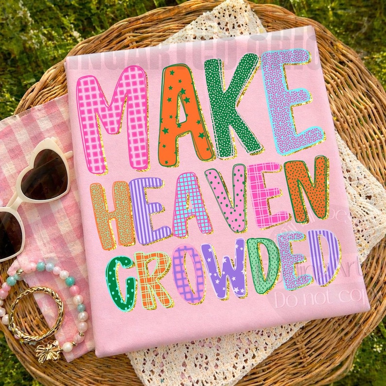 K&ouml;nnte beinhalten: Rosafarbenes T-Shirt mit dem Schriftzug "MAKE HEAVEN CROWDED" in bunten, gemusterten Buchstaben. Das Shirt ist mit verschiedenen Mustern wie Karos, Punkten und Streifen verziert. Das Shirt wird auf einem Weidenkorb mit Sonnenbrille und Schmuck pr&auml;sentiert.