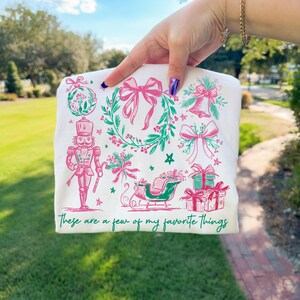 May include: White t-shirt featuring a festive design with pink and green illustrations of Christmas ornaments, bows, a bell, a nutcracker, a sleigh, and wrapped gifts. The text "These are a few of my favorite things" is printed in green.