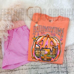 May include: An orange t-shirt featuring a graphic of a jack-o'-lantern with a house inside, the text HALLOWEENTOWN EST. 1998, and stars, paired with pink and white gingham shorts and a gold-toned chain necklace with a circular pendant.