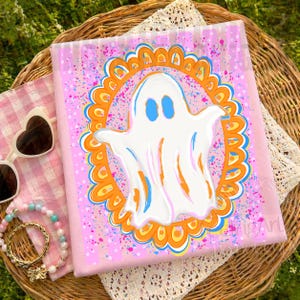 May include: A square pink textile featuring a central white ghost illustration enclosed within an orange and blue scalloped frame. The background is decorated with vibrant pink and blue paint splatters, creating a colourful and festive appearance.