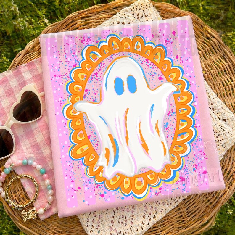 May include: A square pink textile featuring a central white ghost illustration enclosed within an orange and blue scalloped frame. The background is decorated with vibrant pink and blue paint splatters, creating a colourful and festive appearance.