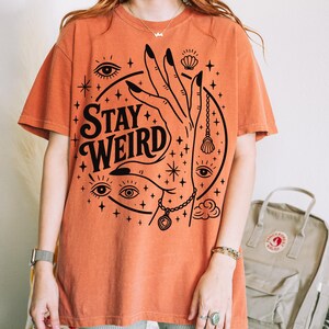 May include: Orange t-shirt with the words "Stay Weird" in a black, stylized font. The graphic includes a hand, eyes, stars, and other mystical symbols. The shirt is a relaxed fit.