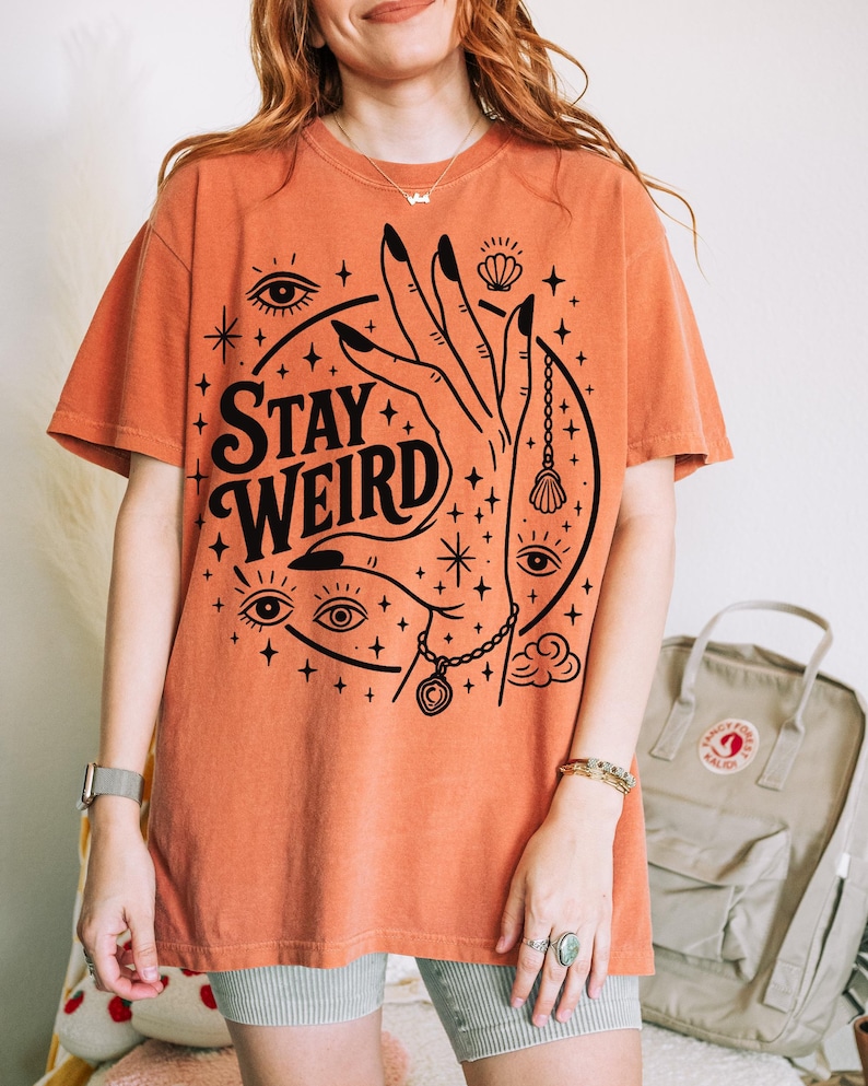 May include: Orange t-shirt with the words "Stay Weird" in a black, stylized font. The graphic includes a hand, eyes, stars, and other mystical symbols. The shirt is a relaxed fit.