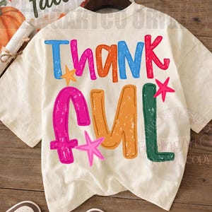 May include: A cream-colored short-sleeve t-shirt featuring the word THANKFUL printed in large, distressed, multi-colored letters. The design includes blue, orange, pink, and green text with small pink and orange star accents on the back.
