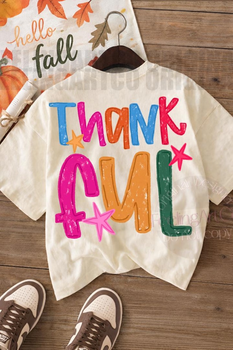 May include: A cream-colored short-sleeve t-shirt featuring the word THANKFUL printed in large, distressed, multi-colored letters. The design includes blue, orange, pink, and green text with small pink and orange star accents on the back.