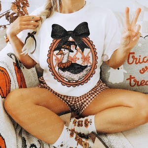 Peut inclure: T-shirt blanc avec un dessin d'oie de dessin anim&eacute; portant un chapeau de sorci&egrave;re et un n&oelig;ud noir. L'oie est dans un cadre ovale avec un motif de n&oelig;uds et d'&eacute;toiles. Le mod&egrave;le porte un short &agrave; carreaux marron et blanc et des chaussettes avec des graphiques d'ours.